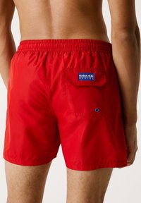 Bañador rojo hecho de un tejido ligero y brillante. Presenta una cintura elástica, un bolsillo trasero con un parche de logo azul y un detalle de ojete.