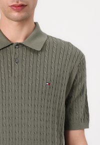Maslinasto zelena polo majica kratkih rukava s uzorkom pletene žice, s izrezanom ovratnikom i dva gumba. Mali logo na prsima.