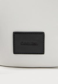 Μαύρη ορθογώνια δερμάτινη ετικέτα Calvin Klein ραμμένη σε ομαλή ανοιχτό γκρι επιφάνεια.