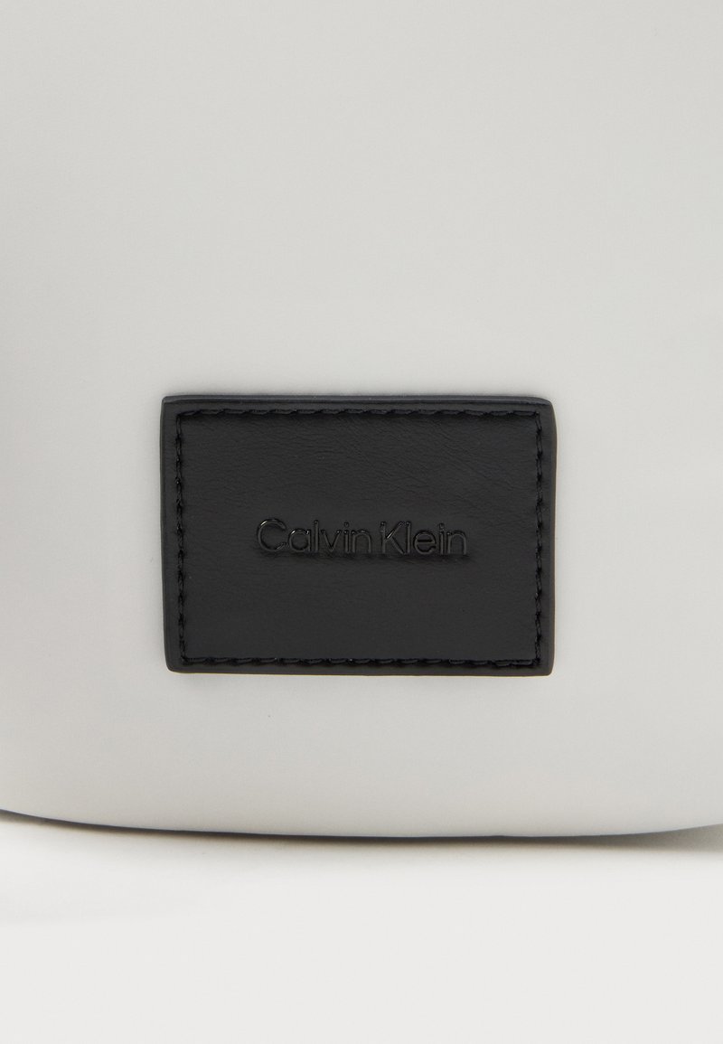 Etiqueta de cuero negra rectangular de Calvin Klein cosida en una superficie lisa de color gris claro.