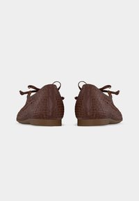 Paire de ballerines en cuir tressé marron avec des bouts ronds et de petits nœuds sur le dessus, vues de dos sur un fond uni.