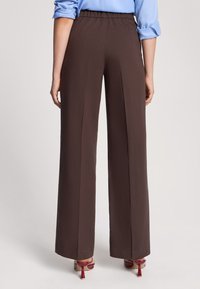 Pantaloni a gamba larga marroni con una texture liscia e vita elastica, abbinati a sandali con tacco rossi. Cuciture a vista e pieghe.