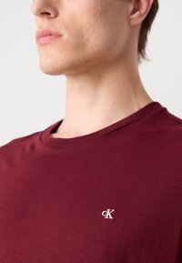 Bordeaux bomulds t-shirt med rund halsudskæring, der har et lille hvidt broderet "CK" logo på venstre side af brystet.