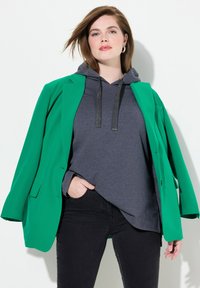 Zelený blazer přes šedou mikinu s kovovými šňůrkami, v kombinaci s černými džíny. Blazer má propracovaný design a jednořadové zapínání.