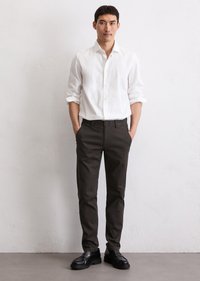 Man stands against white wall wearing white buttoned shirt, black pants, and black shoes with hands in pockets.
