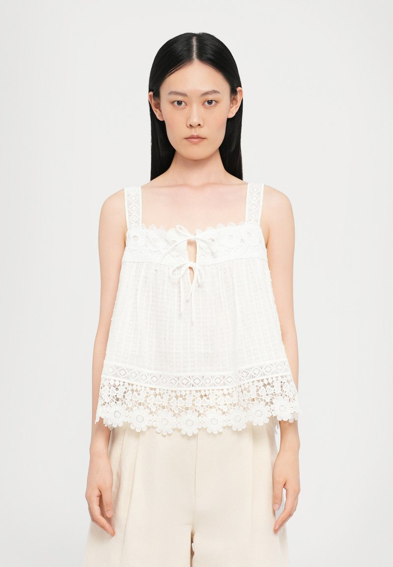 White sleeveless top with a textured check pattern, lace trim, floral accents at the hem, and a front tie detail.