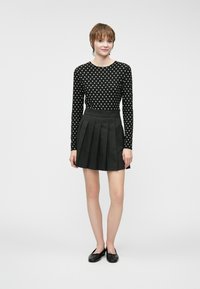 Svart langermet topp med hvite prikker kombinert med et svart plisserte miniskjørt. Modellen har på seg svarte ballettsko. Nøytral bakgrunn.