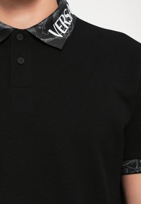 Sort polo shirt med et mønstret krave, der har teksten "VERSACE", to-knaps lukning og tekstureret stof. Kontrasterende kraveaccenter.