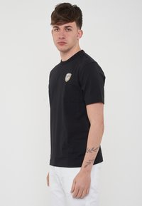 T-shirt in cotone nero a maniche corte, con scollatura rotonda e una piccola grafica a forma di scudo sul lato sinistro. Indossato con pantaloni bianchi.