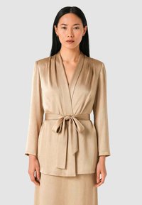 Giacca avvolgente in raso beige con cintura da annodare, maniche lunghe e pieghe verticali sulla parte anteriore, caratterizzata da una texture liscia e una finitura elegante.