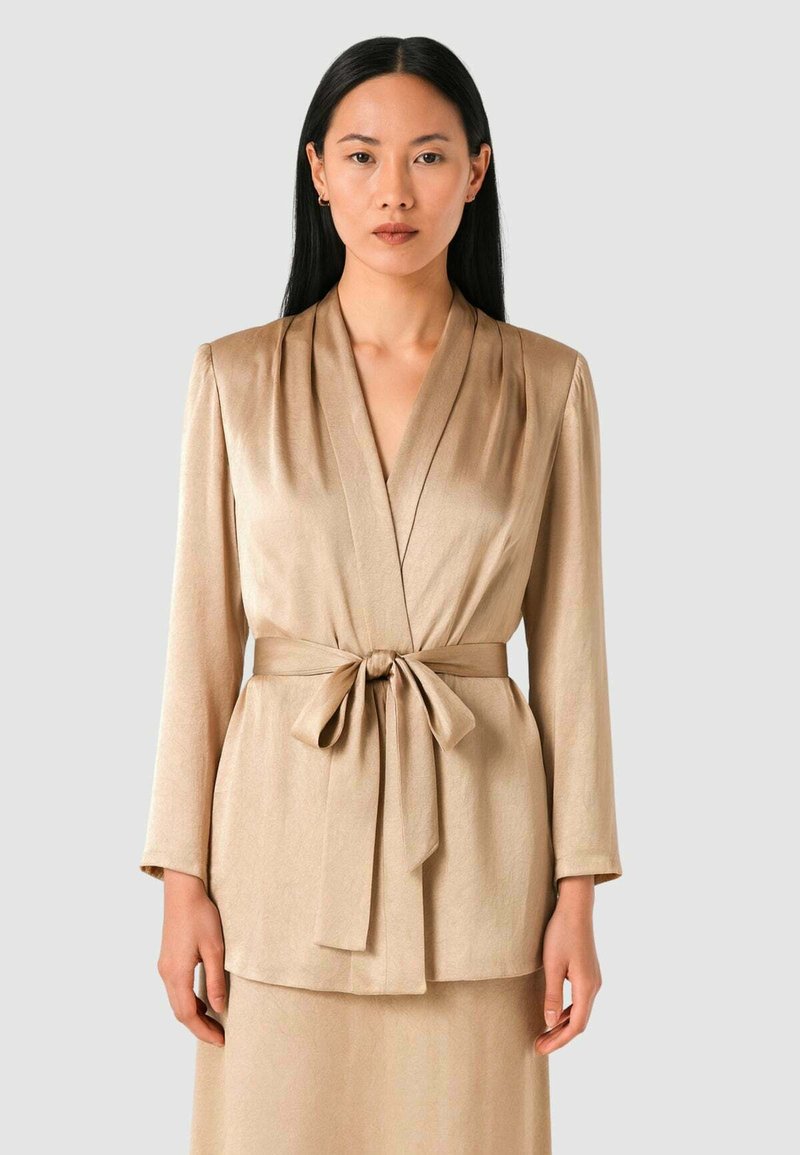 Giacca avvolgente in raso beige con cintura da annodare, maniche lunghe e pieghe verticali sulla parte anteriore, caratterizzata da una texture liscia e una finitura elegante.