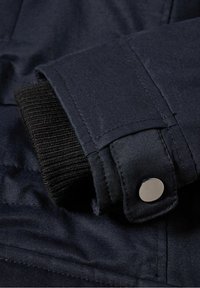 Poignet de veste bleu marine foncé avec un intérieur en maille côtelée noire texturée et un détail de bouton-pression argenté brillant. Texture lisse du tissu extérieur.