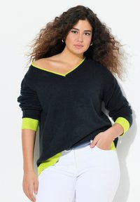 Svart stickad tröja med V-ringning och neongröna detaljer vid kragen och ärmsluten. Bärs med vita jeans med avslappnad passform och textur.