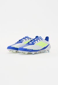 Ghete de fotbal cu un brâu gri deschis, accente albastre și dungi verzi neon. Suprafață texturată cu un profil scăzut și spikes pentru tracțiune.