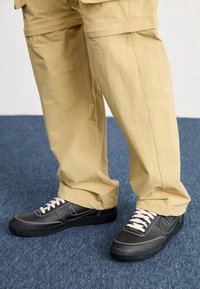Persona que lleva pantalones beige y zapatillas negras con cordones blancos, de pie sobre un suelo de moqueta azul.