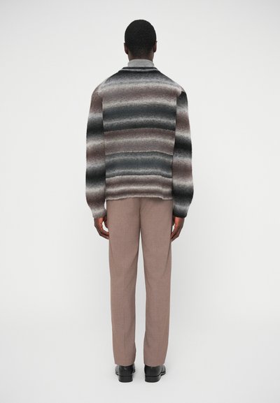 Kootud kampsun horisontaalsete triipudega hallis, pruunis ja mustas; lahtise lõikega; pikad varrukad; kombineeritud helepruunide kohandatud pükstega ja mustade kingadega.