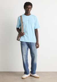 Νεαρός άνδρας με ανοιχτό μπλε T-shirt BOSS, ξεθωριασμένα τζιν με ίσια γραμμή, λευκά αθλητικά παπούτσια και κρατώντας μια μπεζ τσάντα ώμου, που στέκεται σε εσωτερικό χώρο.