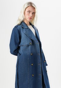 Denim dobbeltradet trenchcoat i dyp blå med notched krage, store knapper og sidelommer. Har en avslappet silhuett.