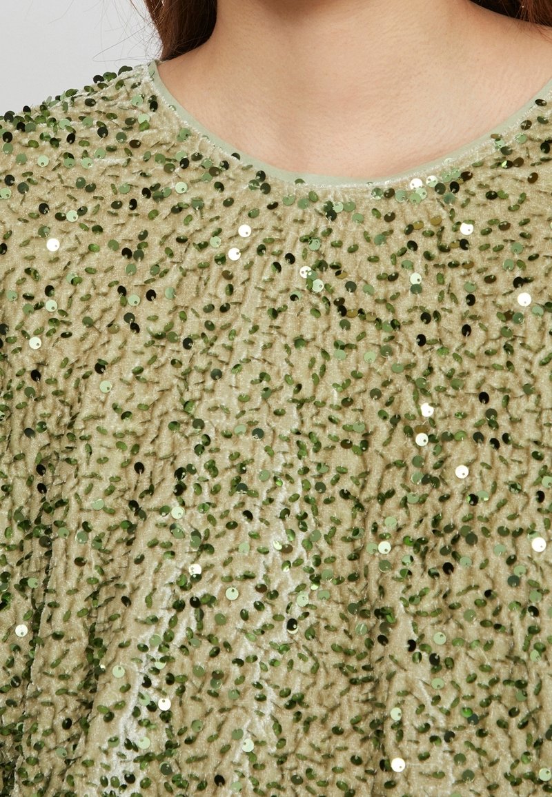 Haut en velours beige à encolure ronde, couvert de sequins verts brillants éparpillés, porté par une personne aux longs cheveux bruns.