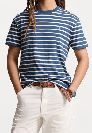 T-shirt rayé bleu et blanc, manches courtes, col rond, tissu en mélange de coton. Porté avec une ceinture tressée marron et un pantalon blanc.