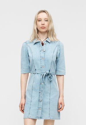 Ung kvinde med glat blondt hår iført en lyseblå denimkjole med knapper og bindebånd i taljen samt en vedhængs halskæde.