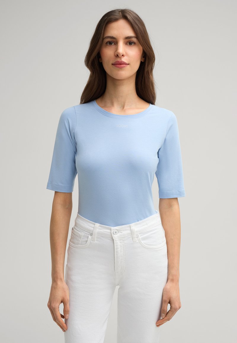 Helles blaues T-Shirt mit kurzen Ärmeln und Rundhalsausschnitt, aus glattem Baumwollstoff gefertigt. Mit dezentem Logo-Stick auf der Brust, kombiniert mit weißen Jeans.