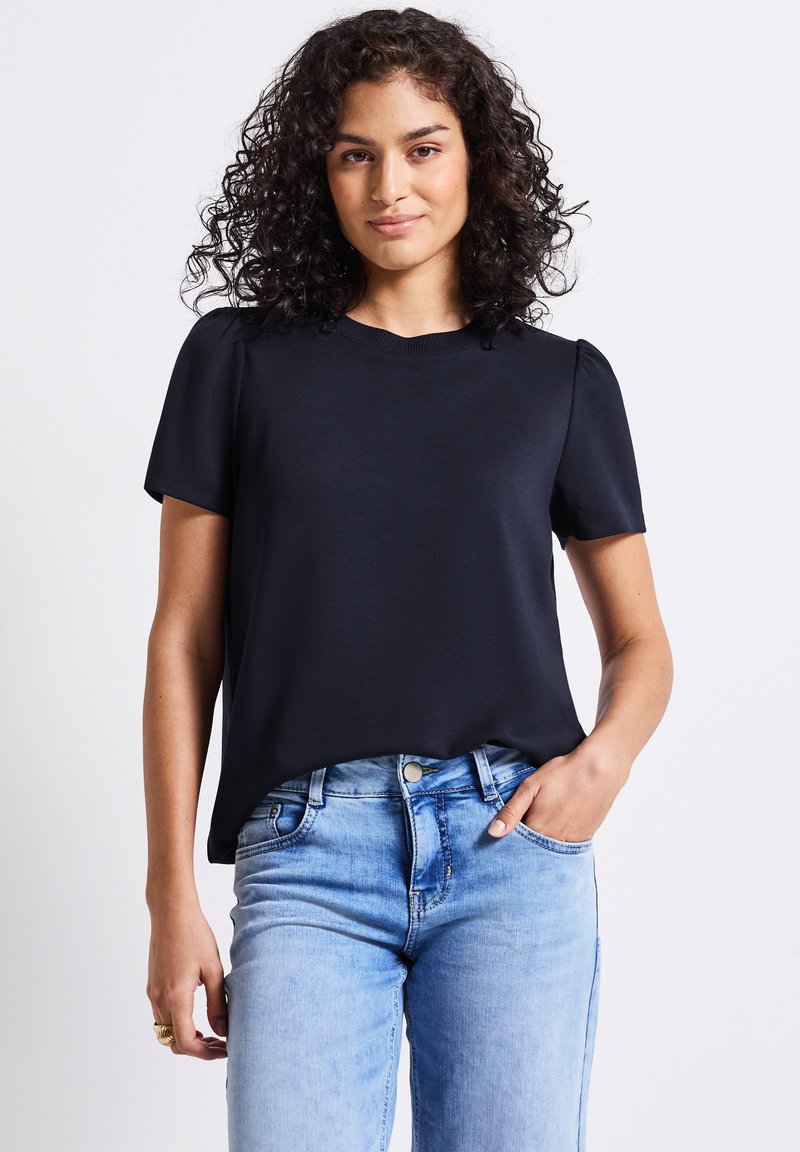 T-shirt noir à manches courtes avec un col rond et une coupe décontractée. Tissu texturé avec des manches légèrement bouffantes. Porté avec un jean bleu clair.