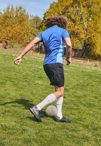 Vêtement de sport bleu et noir avec des manches courtes, short noir et chaussettes blanches jusqu'aux genoux, donnant un coup de pied à un ballon de football de couleur claire sur de l'herbe verte.