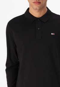 Crna dugmasta polo majica dugih rukava izrađena od teksturiranog materijala, s ovratnikom, tri dugmeta i malim logotipom na lijevom dijelu prsnog koša.