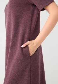 Sukienka w kolorze bordo, wykonana z fakturowanej tkaniny, z krótkimi rękawami, boczną kieszenią i luźnym krojem. Materiał wydaje się miękki i ciepły.