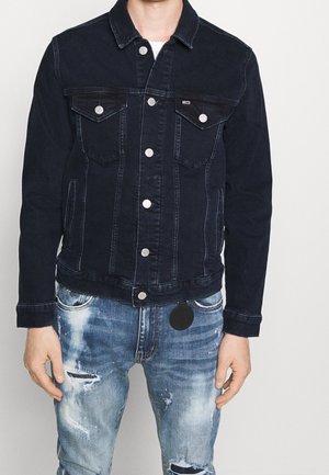 Mężczyzna ubrany w ciemną kurtkę dżinsową zapinaną na guziki oraz zniszczone niebieskie dżinsy z plamami farby i przetarciami.