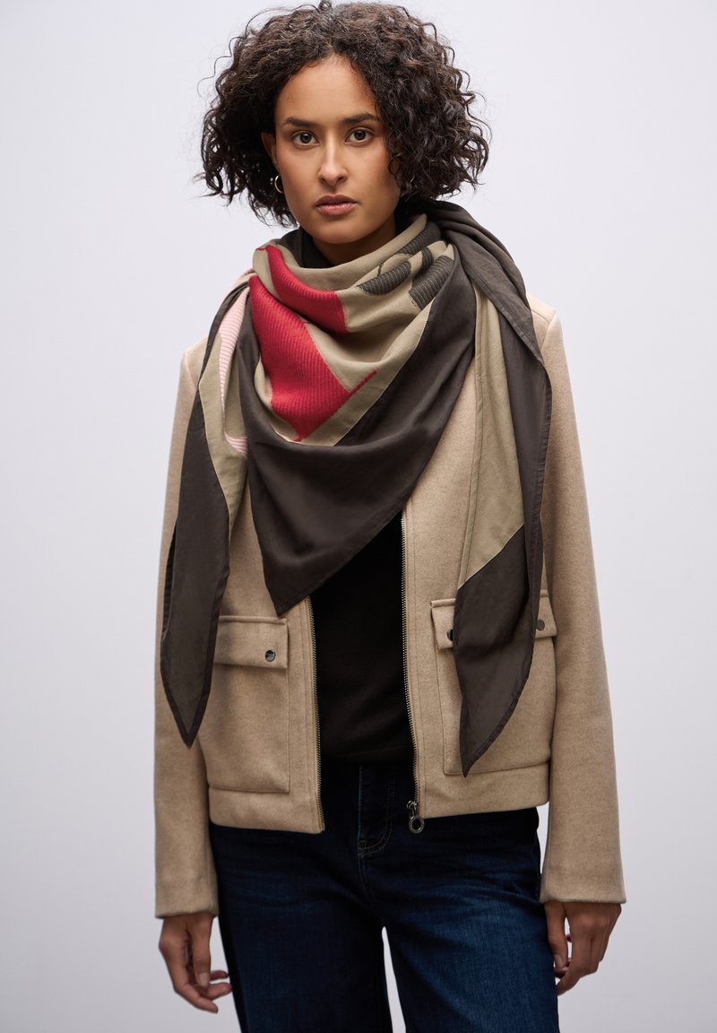 Sciarpa multicolore con motivi geometrici in tessuto rosso, beige e marrone, drappeggiata attorno al collo, abbinata a una giacca beige.