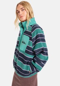 Fleece pulover jakna u tirkiznoj i tamnoplavoj boji, s horizontalnim prugama i džepom sa zatvaračem. Tekstuirana tkanina i ovratnik koji stoji.