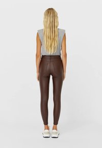 Vrouw met lang blond haar in een grijze mouwloze top, strakke bruine leren broek en witte sneakers, staande met haar rug naar de kijker.