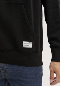 Sudadera negra con un bolsillo frontal y puños acanalados. Presenta una etiqueta de tela con el nombre de la marca y las instrucciones de cuidado. Combinada con unos jeans azules.
