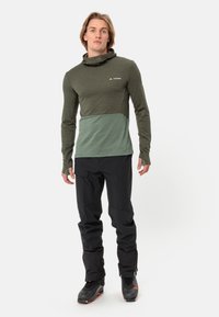 Grünes Langarmshirt mit einem helleren grünen unteren Bereich, kombiniert mit schwarzen Hosen und Stiefeln, ausgestattet mit einer Kapuze und Daumenlöchern.