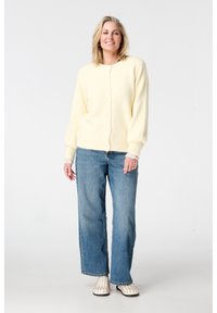 Lichtgeel, zacht vest met een ronde hals en knopen, gecombineerd met blauwe jeans met brede pijpen en crèmekleurige instapschoenen met studs.