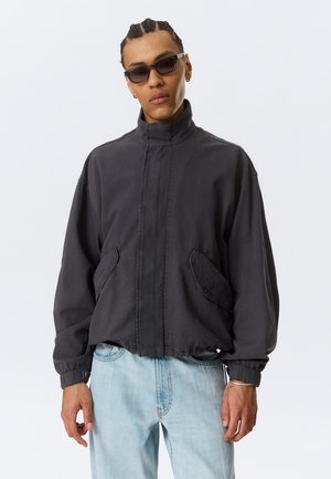 Jeune homme aux cheveux nattés portant une veste sombre oversized, un jean bleu clair, des lunettes de soleil et de petites créoles sur un fond uni.