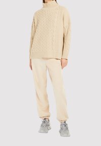 Beige jättikokoinen neulepusero korkealla kauluksella ja letinukkekuvioilla, yhdistetty vastaaviin beigeihin vapaa-ajan housuihin ja vaaleanharmaisiin lenkkareihin.