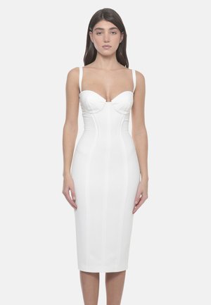 Donna con i capelli lunghi e scuri che indossa un abito midi bianco aderente con spalline sottili e dettagli strutturati sul busto, in piedi contro uno sfondo semplice.