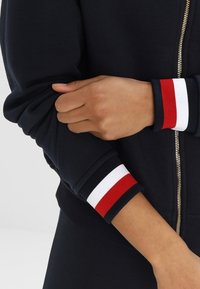 Tmavě modrá bunda na zip s žebrovanými manžetami lemovanými červenými, bílými a tmavě modrými pruhy. Detailní pohled na paži držící rukáv. Hladká textura.