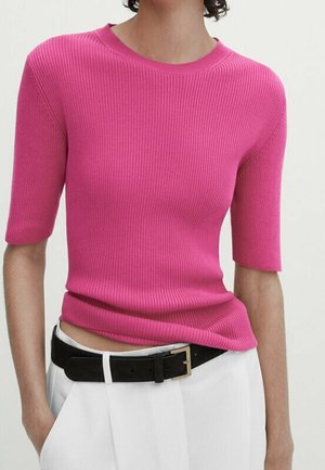 Woman wearing a fitted, ribbed, bright pink short-sleeve top tucked into white trousers with a black belt.