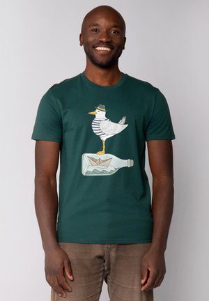Green cotton t-shirt featuring a graphic of a seagull in a striped shirt standing on a bottle with a paper boat inside.