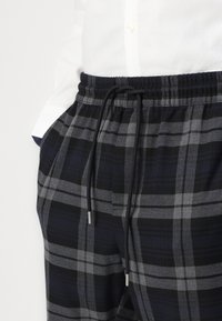 Zwarte en grijze geruite broek met een elastische tailleband en trekkoord. Beschikt over metalen uiteinden aan het trekkoord en een zachte stoftextuur.