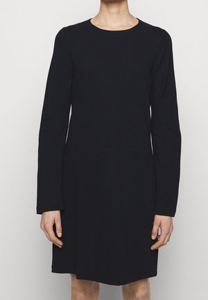 Person wearing a simple, long-sleeved black dress with a round neckline, standing against a plain light background.