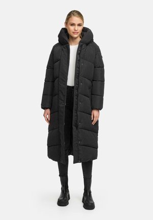 Long black puffer coat with a high collar, quilted design, snap closures, and side pockets. Worn over a white top and black pants.