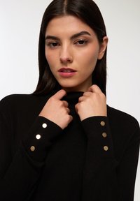 Czarny sweter z prążkowanego materiału z wysokim kołnierzem i trzema złotymi guzikami na każdym mankiecie. Materiał wydaje się miękki o gładkiej teksturze.