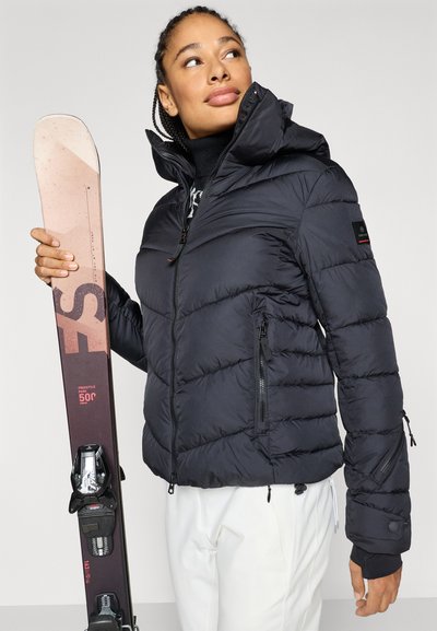 Casaco acolchoado preto com gola alta, bolsos com fecho e punhos elásticos; segurando esquis bege com fixações pretas, contra um fundo claro.