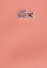 Rosa texturerad tyg med en broderad krokodillogo med vit kontur och röda detaljer.