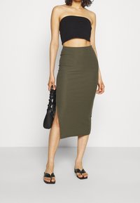 Žena oblečená v černém topu bez ramínek, olivově zelené midi sukni s žebrovaným vzorem a bočním rozparkem, černé zmačkané kabelce a černých sandálech na otevřenou špičku.